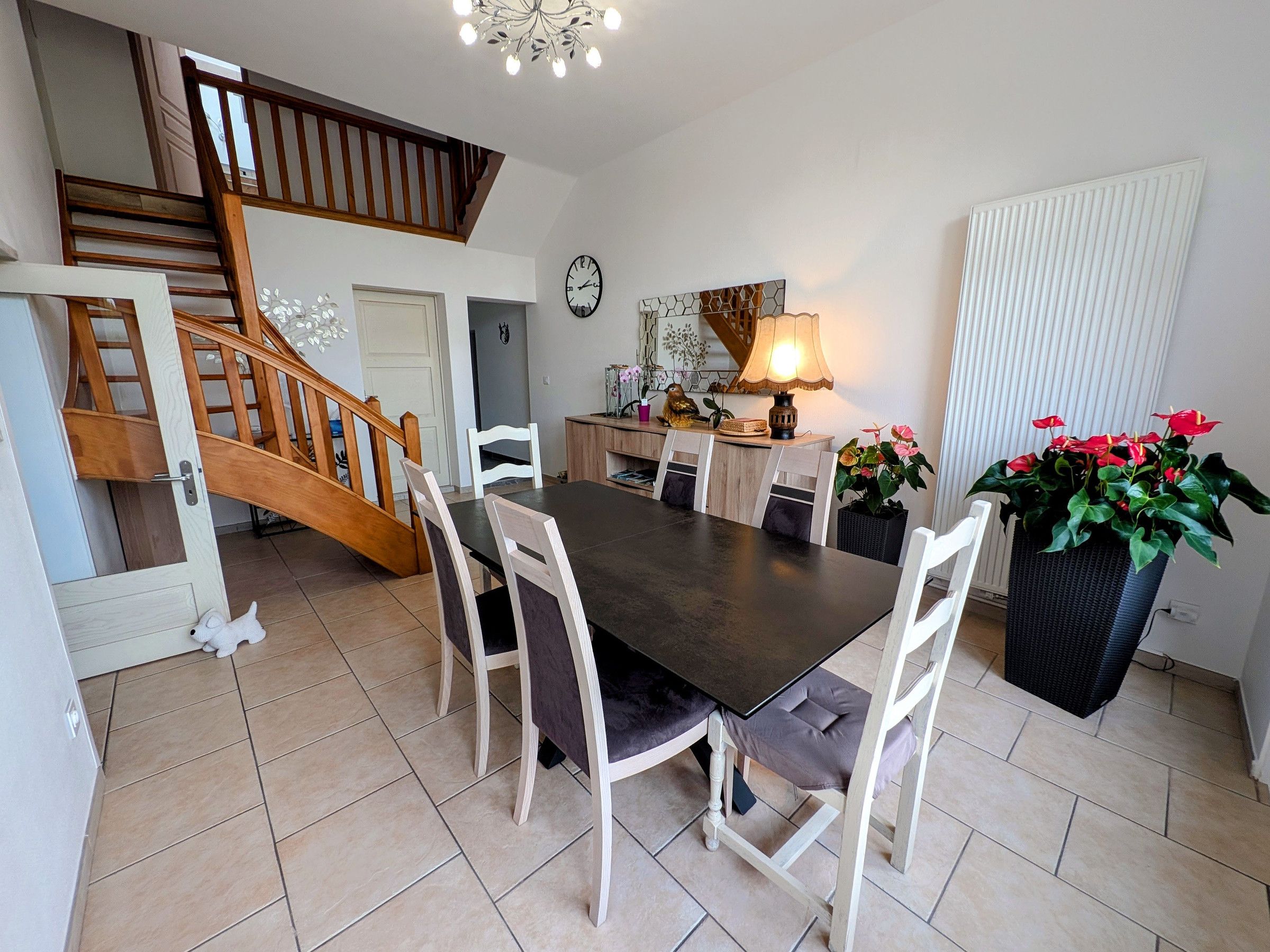 Un coin repas avec une table en bois foncé, des chaises blanches, un escalier, une horloge murale, une lampe et une plante en pot.