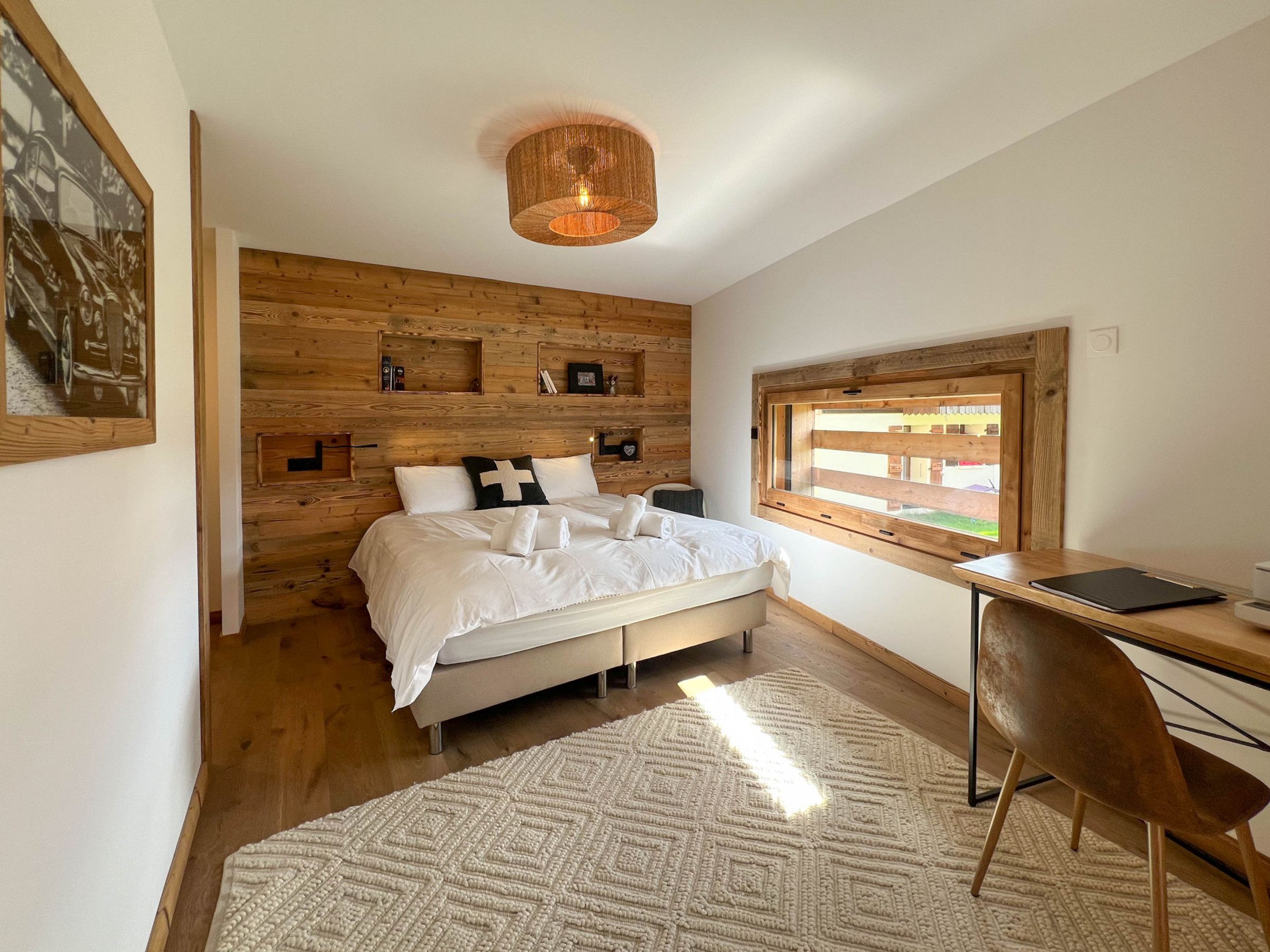 Une chambre à coucher avec une tête de lit en bois, un lit avec une literie blanche, un bureau avec une chaise et une fenêtre donnant sur un paysage agréable.