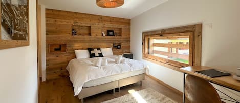 Un dormitorio con cabecera de madera, cama con ropa de cama blanca, escritorio con silla y ventana con vistas.