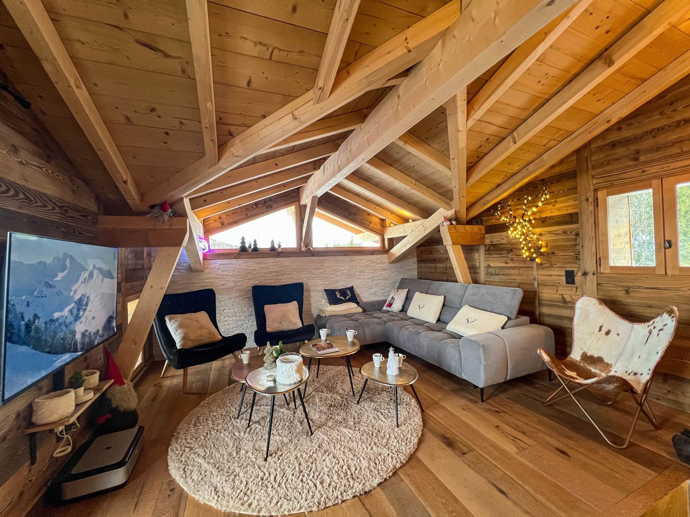 Ein gemütliches Blockhaus-Interieur mit Schlafgalerie, einem Wohnbereich inklusive Sofa und Sesseln sowie einem Fernseher, der einen Blick auf die Berge zeigt.