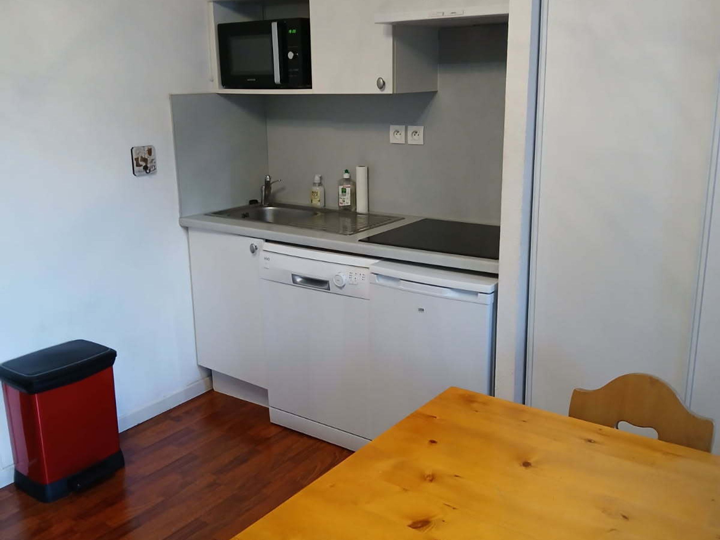 A compact kitchen with a microwave, sink, and dishwasher. There's a wooden table and a trash bin.