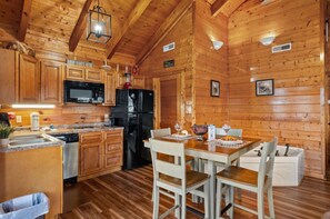 A wooden cabin interior with a kitchen featuring a microwave, refrigerator, and wooden cabinets. There is a dining table with chairs and a small trash bin. - Little River Cabin | Studio Log Cabin Near Downtown Pigeon Forge (Pigeon Forge)