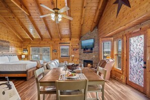 A cozy cabin interior with a wooden ceiling, a fireplace, a dining table with chairs, a bed, and a television mounted on the wall. - Little River Cabin | Studio Log Cabin Near Downtown Pigeon Forge (Pigeon Forge)