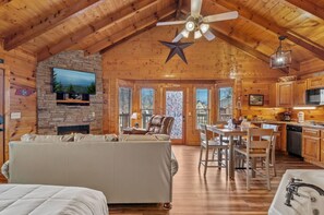 A cozy log cabin interior with a stone fireplace, a large star ceiling fan, a dining area, and a kitchen with wooden cabinets. - Little River Cabin | Studio Log Cabin Near Downtown Pigeon Forge (Pigeon Forge)