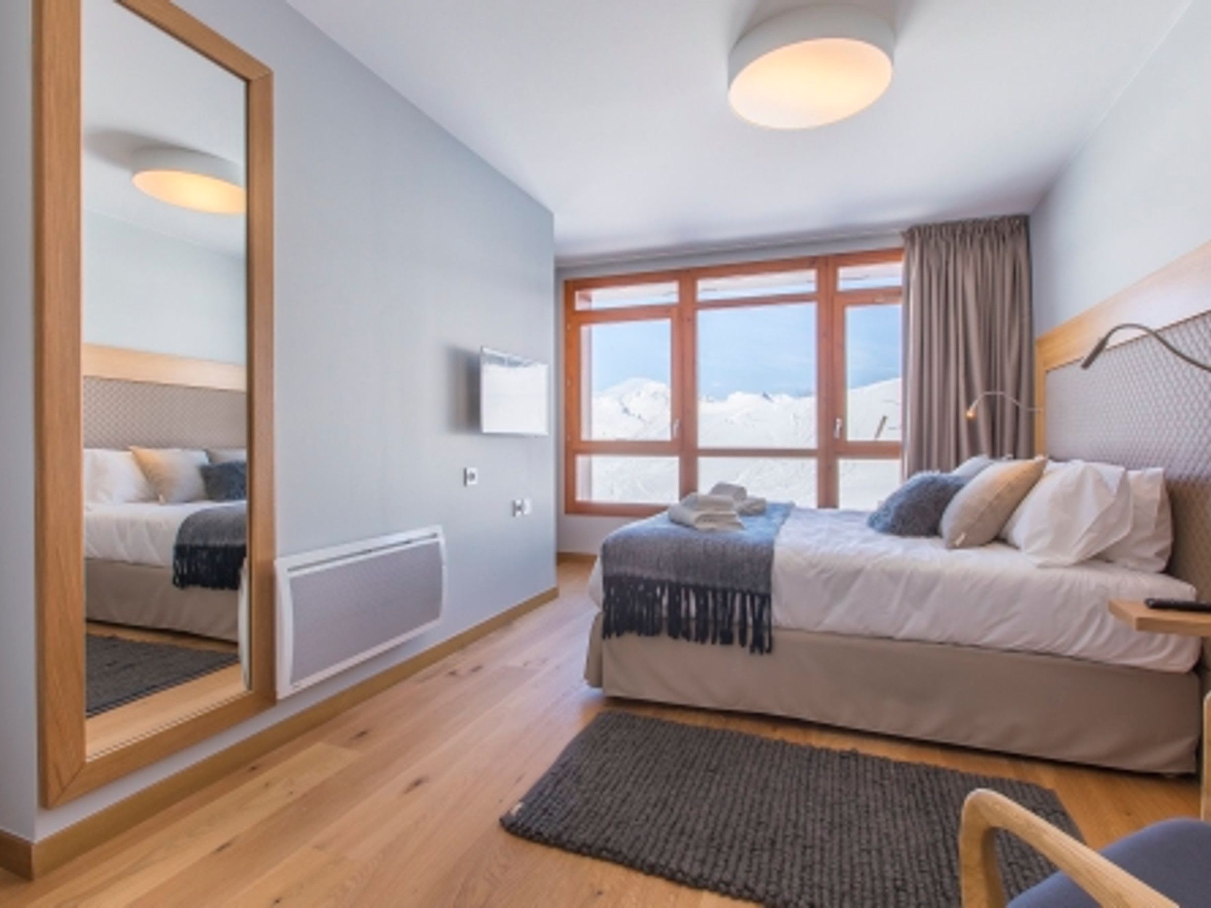 Een slaapkamer met een grote spiegel, een bed met hoofdbord, een raam met gordijnen en uitzicht op besneeuwde bergen.