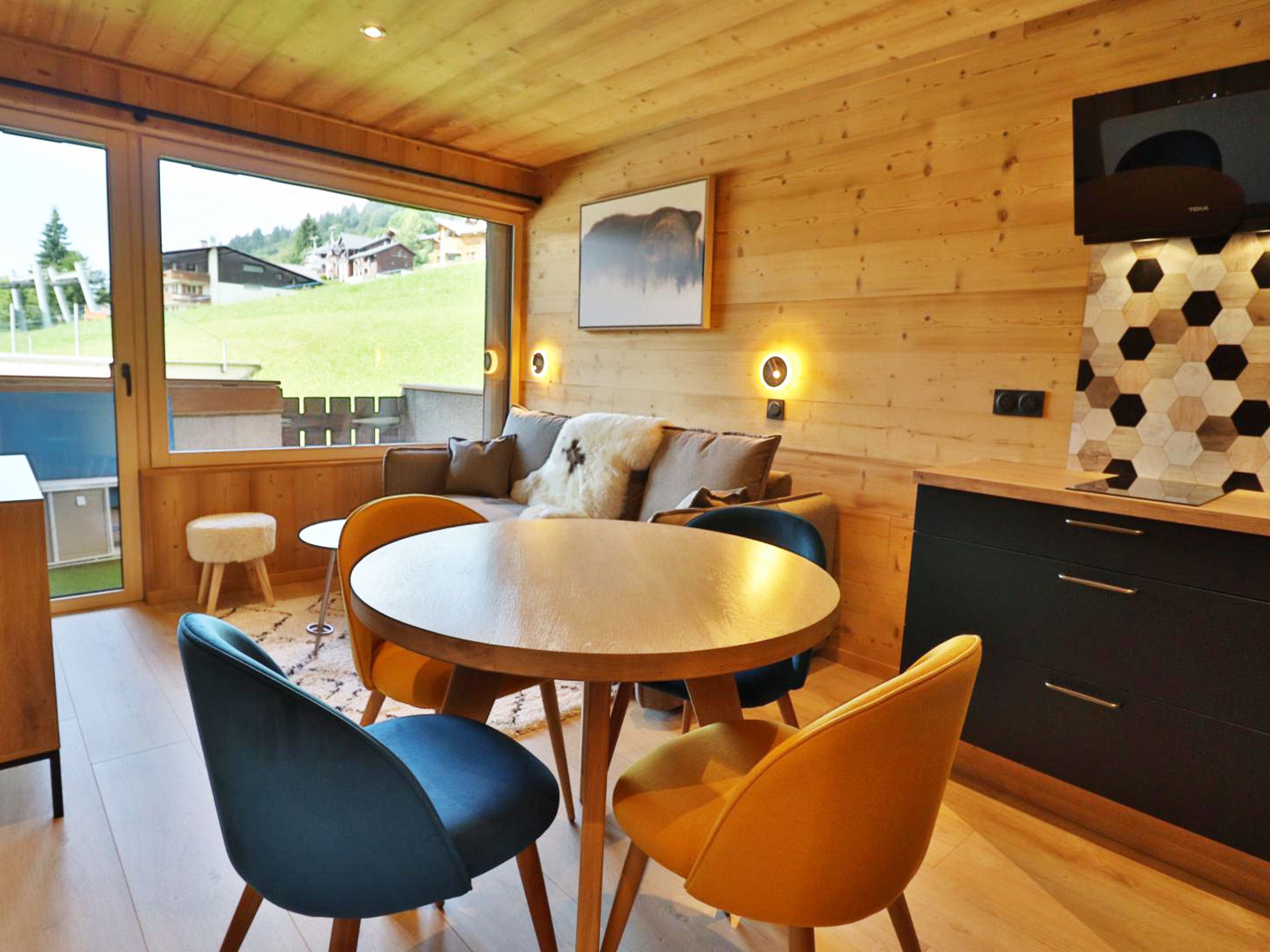 Un acogedor interior de cabaña con una mesa redonda de comedor, sillas de madera, un sofá y un televisor montado en la pared.