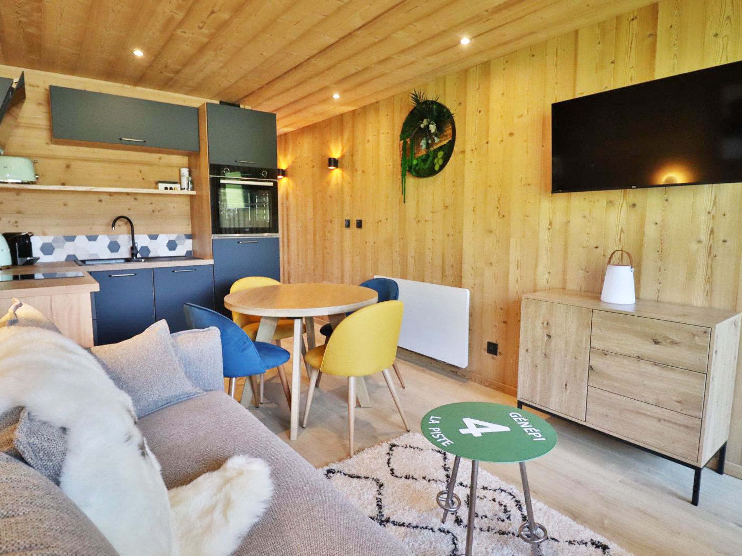 Quarto estilo cabana aconchegante com sofá, mesa de jantar, cadeiras, cozinha compacta e TV na parede.