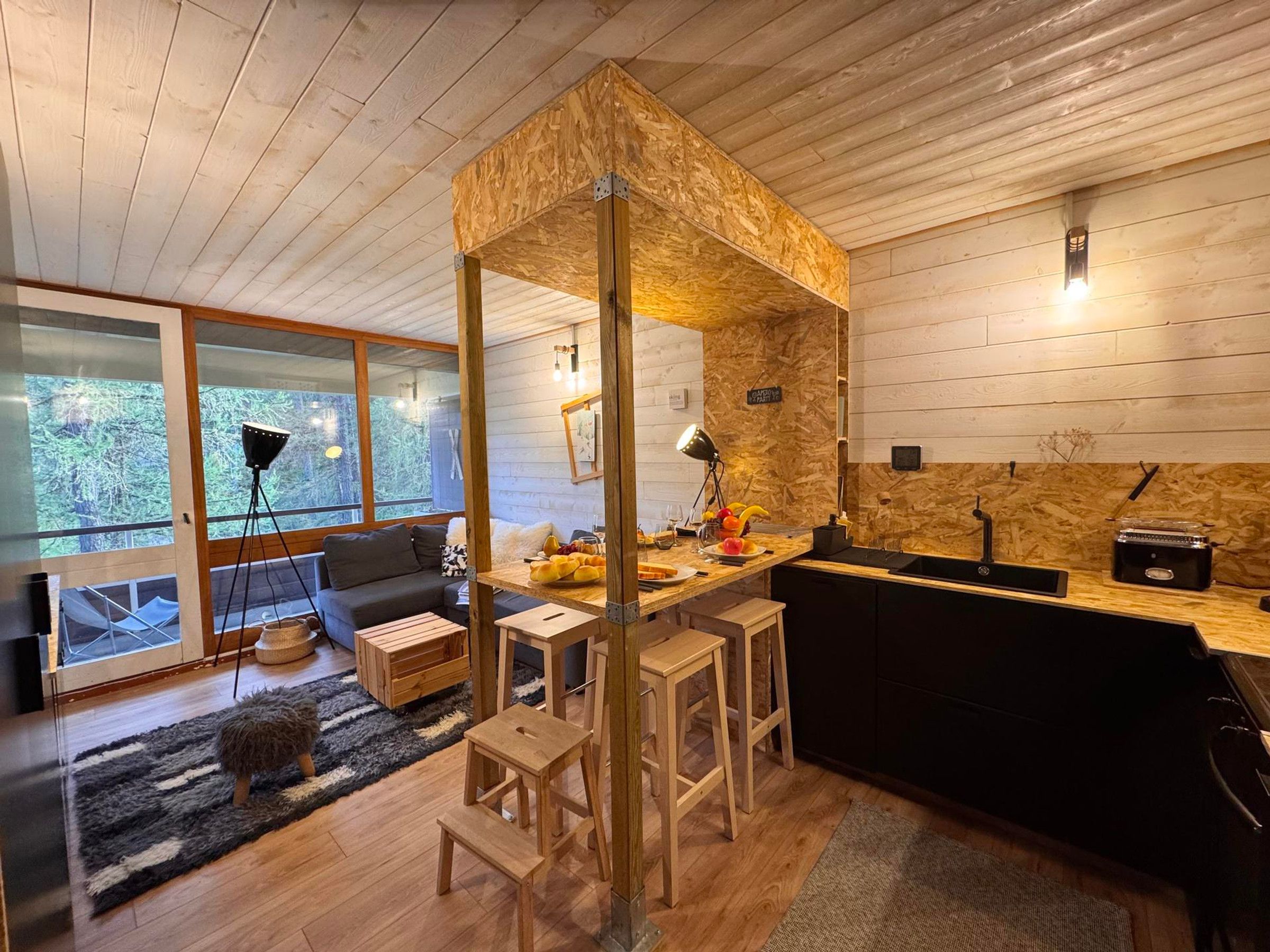 Cozinha moderna com detalhes em madeira, área de refeições com sofá e uma grande janela com vista para árvores.