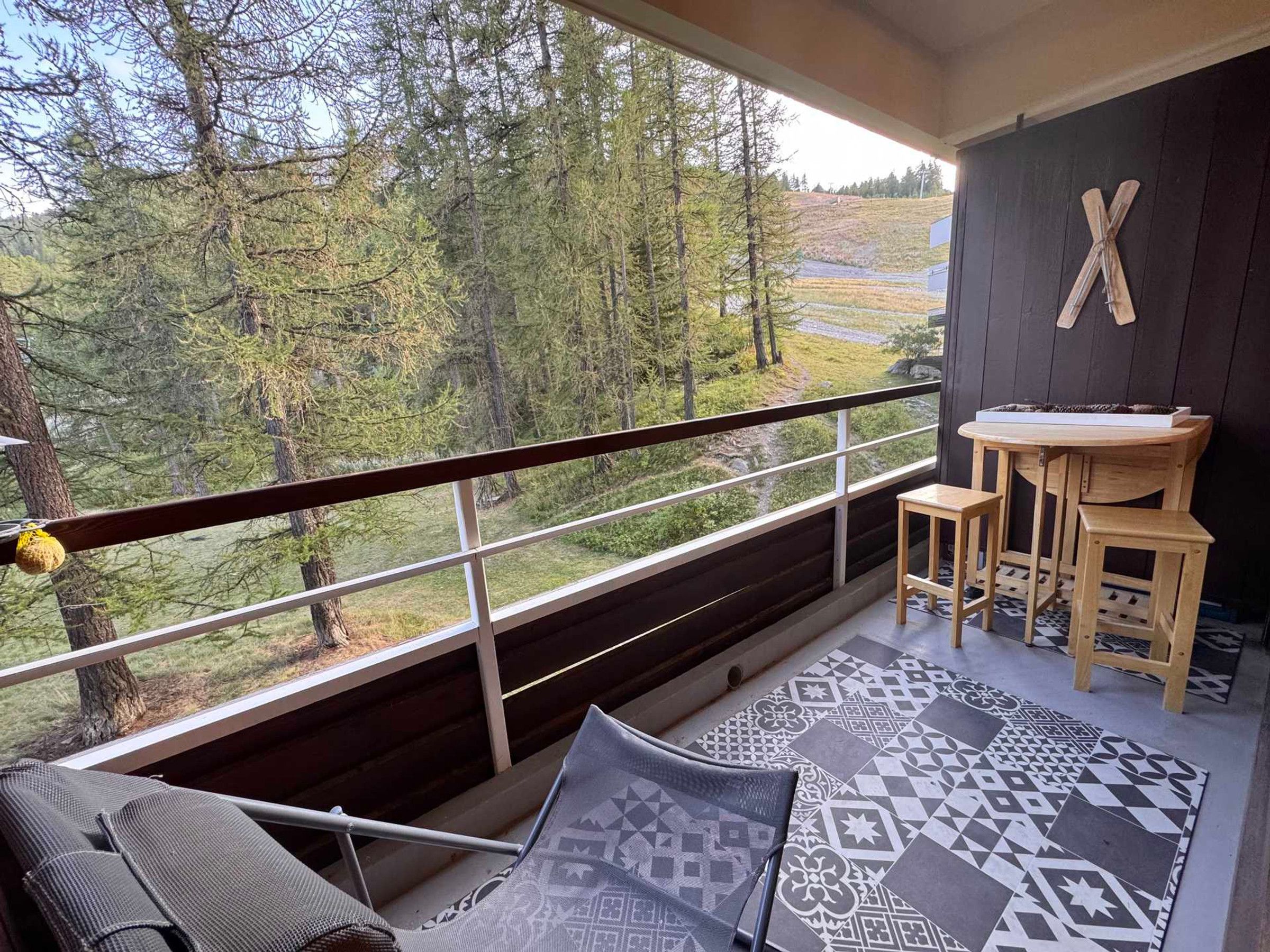 A balcony with a view of a forest, a wooden table with two chairs, and a patterned rug.
