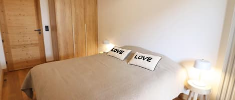 A bedroom with a bed, two pillows with the word 'LOVE,' a nightstand with a lamp, a wooden wardrobe, and a door.