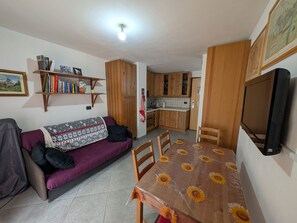 Un espacio habitable compacto con un sofá, una mesa de comedor con mantel de girasoles, armarios de madera, un televisor y una estantería con libros.