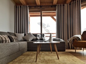 A modern living room with a sofa, a coffee table, and a chair. There is a window with curtains, and a view of a snowy landscape. - Exceptional apartment, mountain view - 10 guests (Les Deux Alpes)