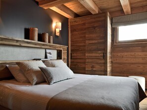 A bedroom with a wooden headboard, a bed with pillows, a nightstand, a wall-mounted lamp, and a window. - Exceptional apartment, mountain view - 10 guests (Les Deux Alpes)