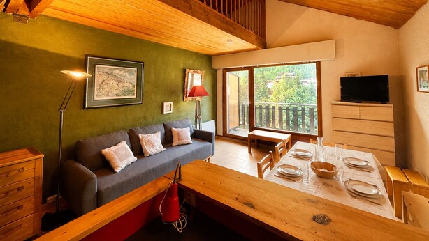 Ein gemütliches Wohnzimmer mit Holzbalkendecke, einer Couch mit Kissen, einem Esstisch für vier Personen und einem Fernseher an der Wand.