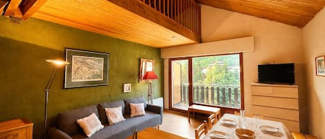 A cozy living room with a wooden ceiling, a sofa with cushions, a dining table set for four, and a television on the wall.