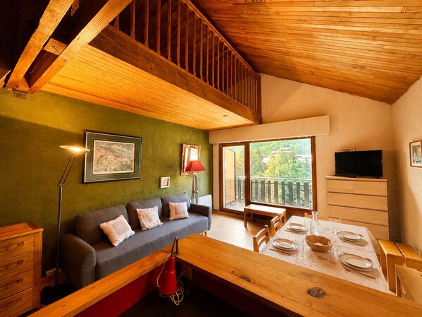 A cozy living room with a wooden ceiling, a sofa with cushions, a dining table set for four, and a television on the wall. - Central T1 loft with parking - 4 guests (Les Deux Alpes)