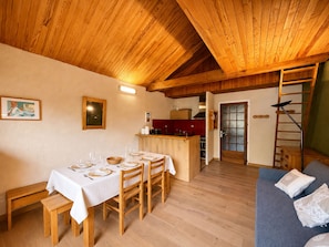A dining area with a wooden table set for four, a kitchen in the background, and a staircase leading to an upper level. - Central T1 loft with parking - 4 guests (Les Deux Alpes)