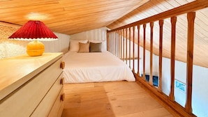 Ein Schlafzimmer mit geneigter Decke, Holzboden, einem Bett mit weißer Bettwäsche, einem roten Lampenschirm und einem Holzgeländer.