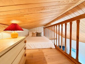 A bedroom with a sloped ceiling, wooden flooring, a bed with white bedding, a red lampshade, and a wooden railing. - Central T1 loft with parking - 4 guests (Les Deux Alpes)