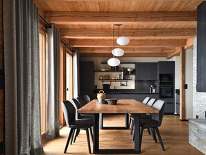Ein moderner Essbereich mit Holztisch und -stühlen, im Hintergrund eine Küche und ein großes Fenster mit Vorhängen.