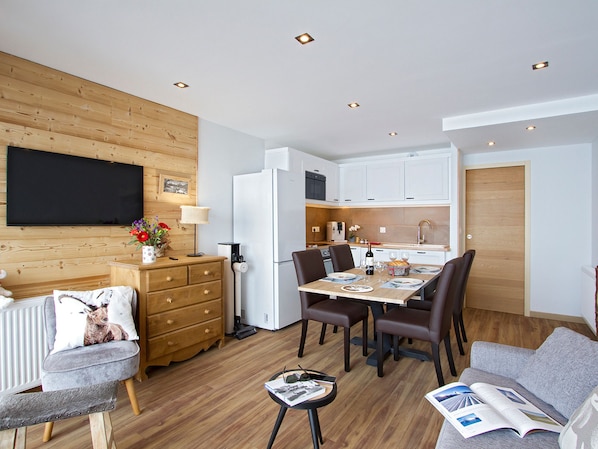 A modern kitchen with white cabinets, a dining area with a table set for four, and a television mounted on a wooden wall. - Apartment with view of La Muzelle - 4 guests (Les Deux Alpes)