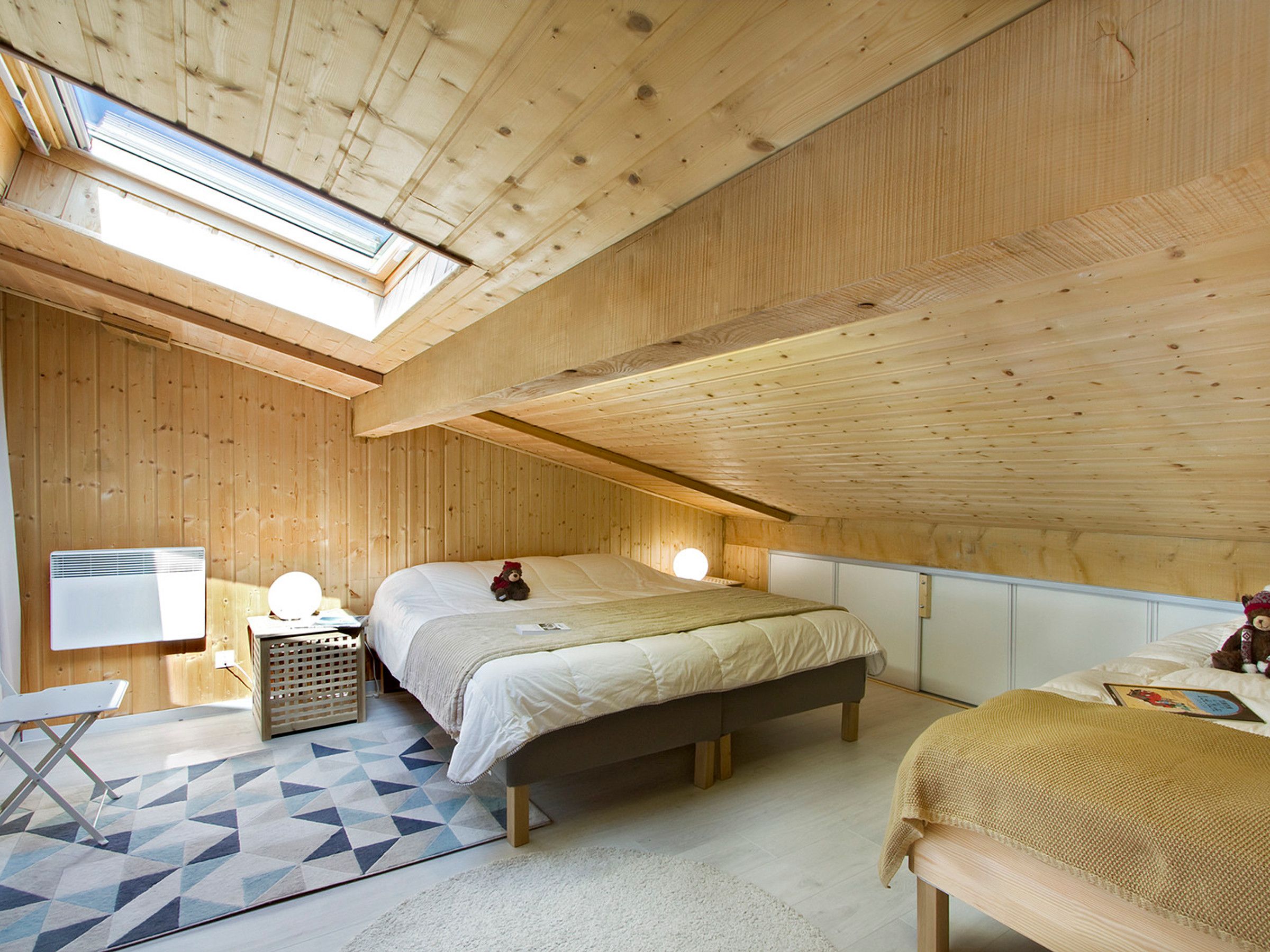Ein Schlafzimmer mit schrägem Dach, einem Bett, Nachttischlampen, einem kleinen Tisch und einem Sessel.