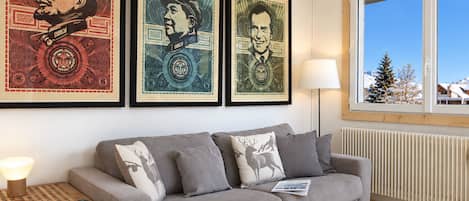 Un salon avec un canapé gris, une table d’appoint en bois sur laquelle se trouve une lampe, et trois tableaux encadrés accrochés au mur.