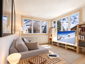 A living room with a sofa, a coffee table with a chess set, a TV displaying a snowy landscape, and a bookshelf. - Cozy duplex, near slopes - 8 guests (Les Deux Alpes)