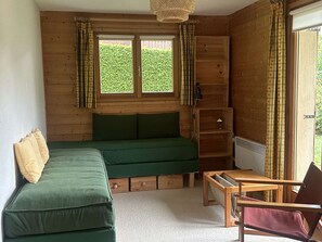 Ein gemütliches Zimmer mit einem grünen Sofa, einem Holztisch und einem Stuhl. Es gibt Fenster mit Vorhängen, ein Regal und eine Heizung.