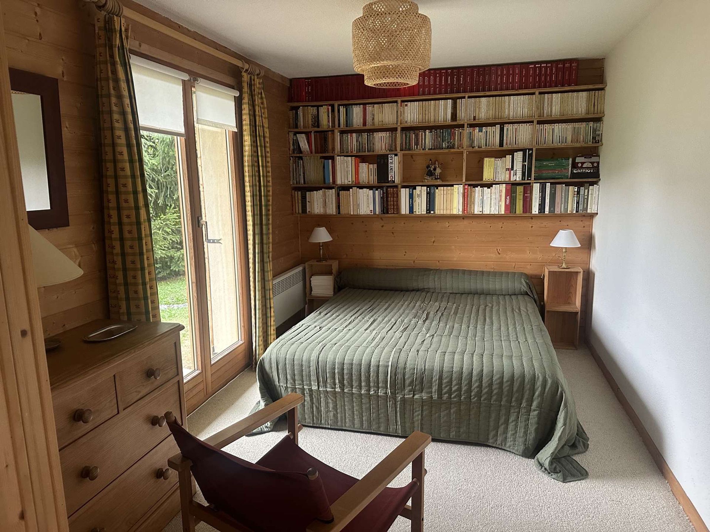 A bedroom with a bed, a wooden dresser, a chair, a bookshelf, and a window with curtains.