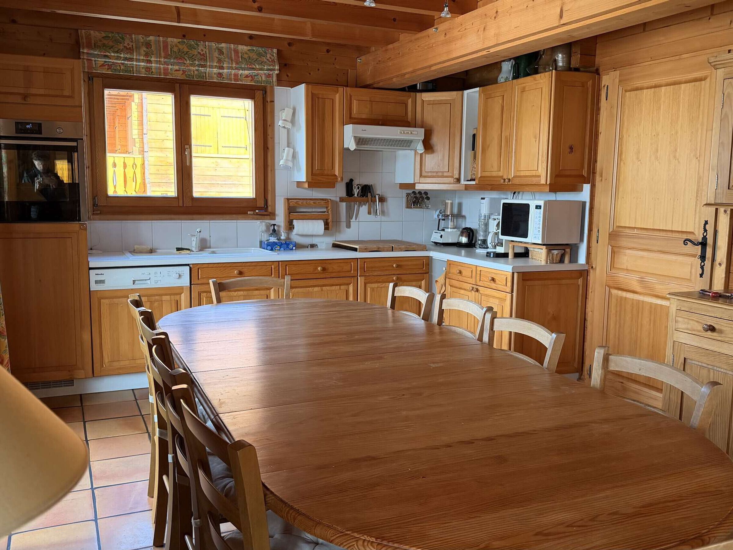 Una cocina de madera con una mesa de comedor y sillas, electrodomésticos blancos y un ventanal con cortinas.