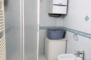 Un bagno con un lavandino bianco, un portascopino, una bacinella di plastica blu e un water bianco.