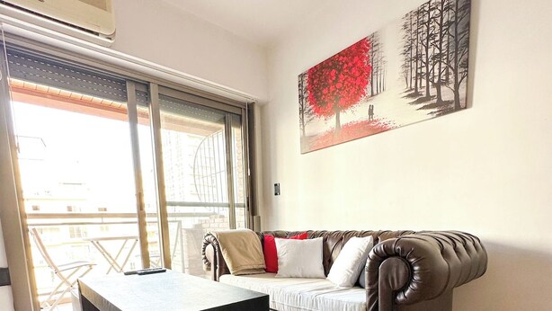 A living room with a sofa, a coffee table, and a small ottoman. There is a painting on the wall and a window with a view of buildings. - Three bedrooms with parking and security. (Buenos Aires)