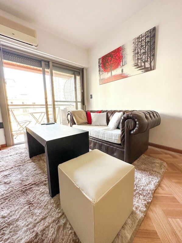 A living room with a sofa, a coffee table, and a small ottoman. There is a painting on the wall and a window with a view of buildings. - Three bedrooms with parking and security. (Buenos Aires)