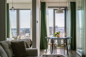 Un salon moderne avec un canapé, une table à manger et des chaises, ainsi que de grandes fenêtres ornées de rideaux verts.