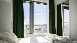 A hotel room with a large bed, a view of a cityscape through two windows, and a neatly made bed with a towel. - Kalasatama apartments by Come in (Helsinki)