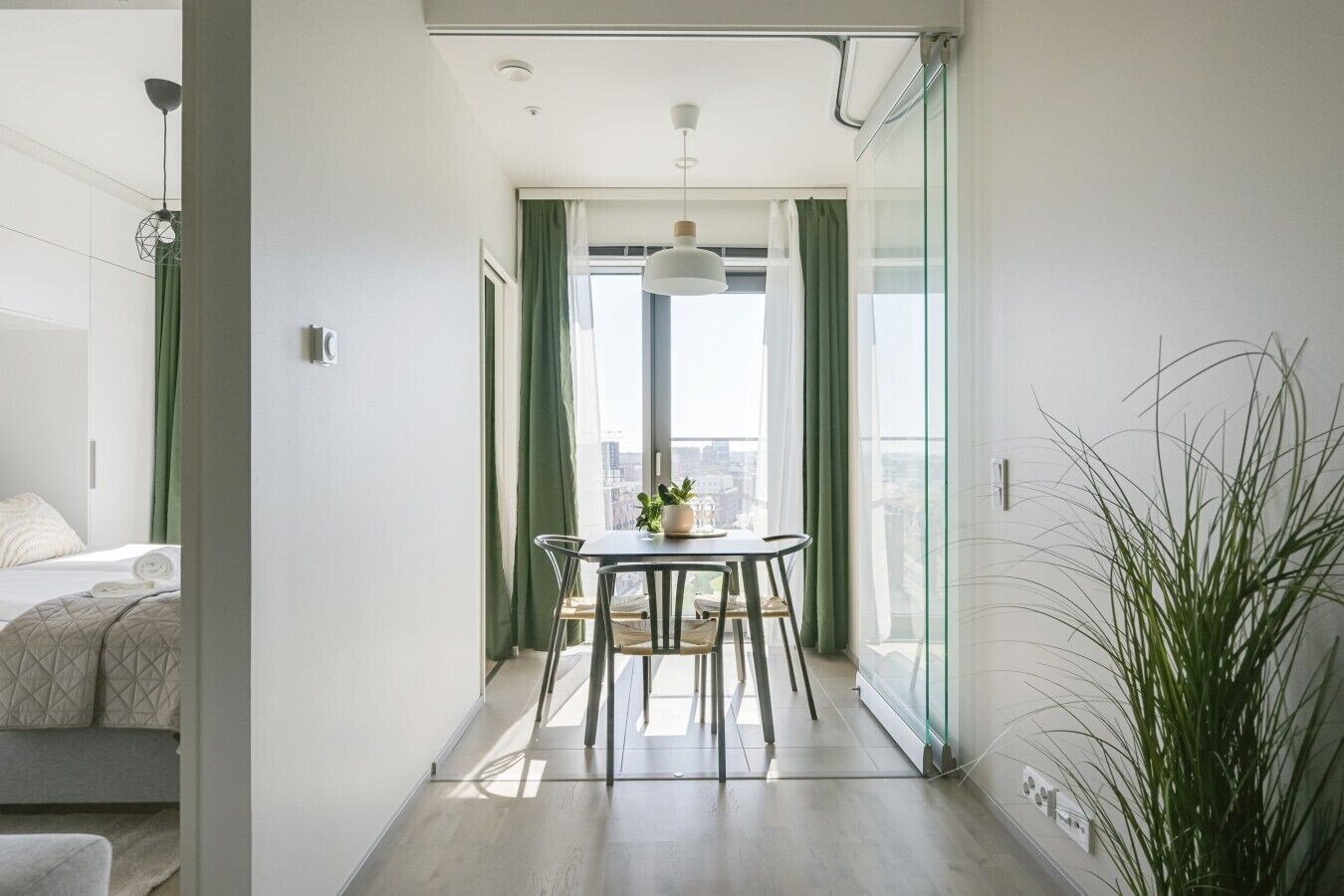 Habitación de hotel moderna con balcón, zona de comedor con mesa y sillas, y cama con cortina verde.