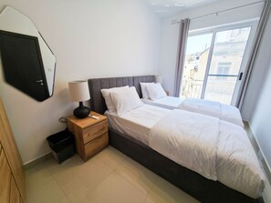 Ein Schlafzimmer mit zwei Betten, einem Nachttisch, einem Spiegel und Blick auf Gebäude durch das Fenster.