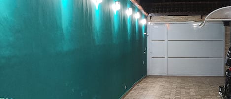 A covered outdoor area with a tiled floor, a green wall with lights, and a white gate.
