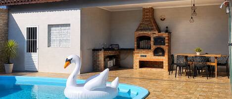 A swimming pool with a swan float, a brick barbecue area, and a patio with chairs.