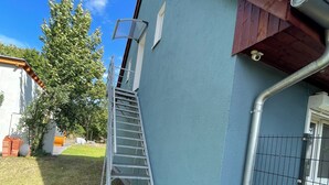 A building with a metal staircase leading to a door, a fenced area, and a tree in the background. - Ferienwohnung für 4 Personen in Plattenburg (Plattenburg)