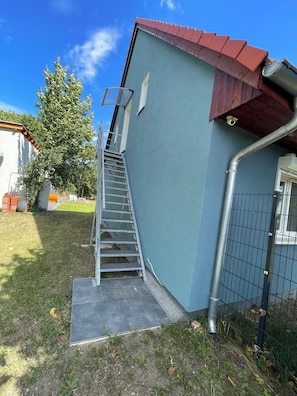 A building with a metal staircase leading to a door, a fenced area, and a tree in the background. - Ferienwohnung für 4 Personen in Plattenburg (Plattenburg)