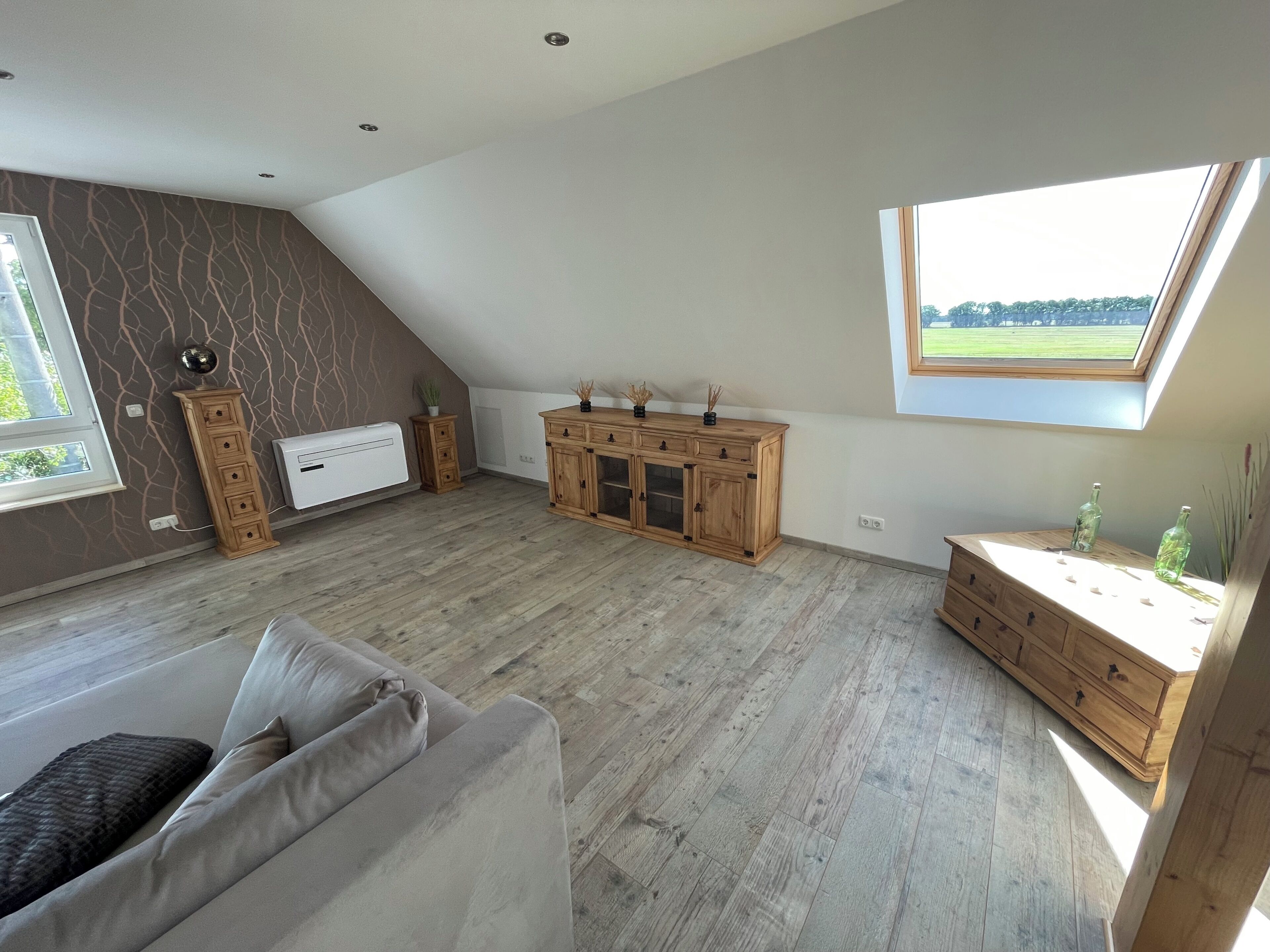 A spacious loft bedroom with a sloped ceiling, a built-in wardrobe, a large window with a view, and a grey sofa.