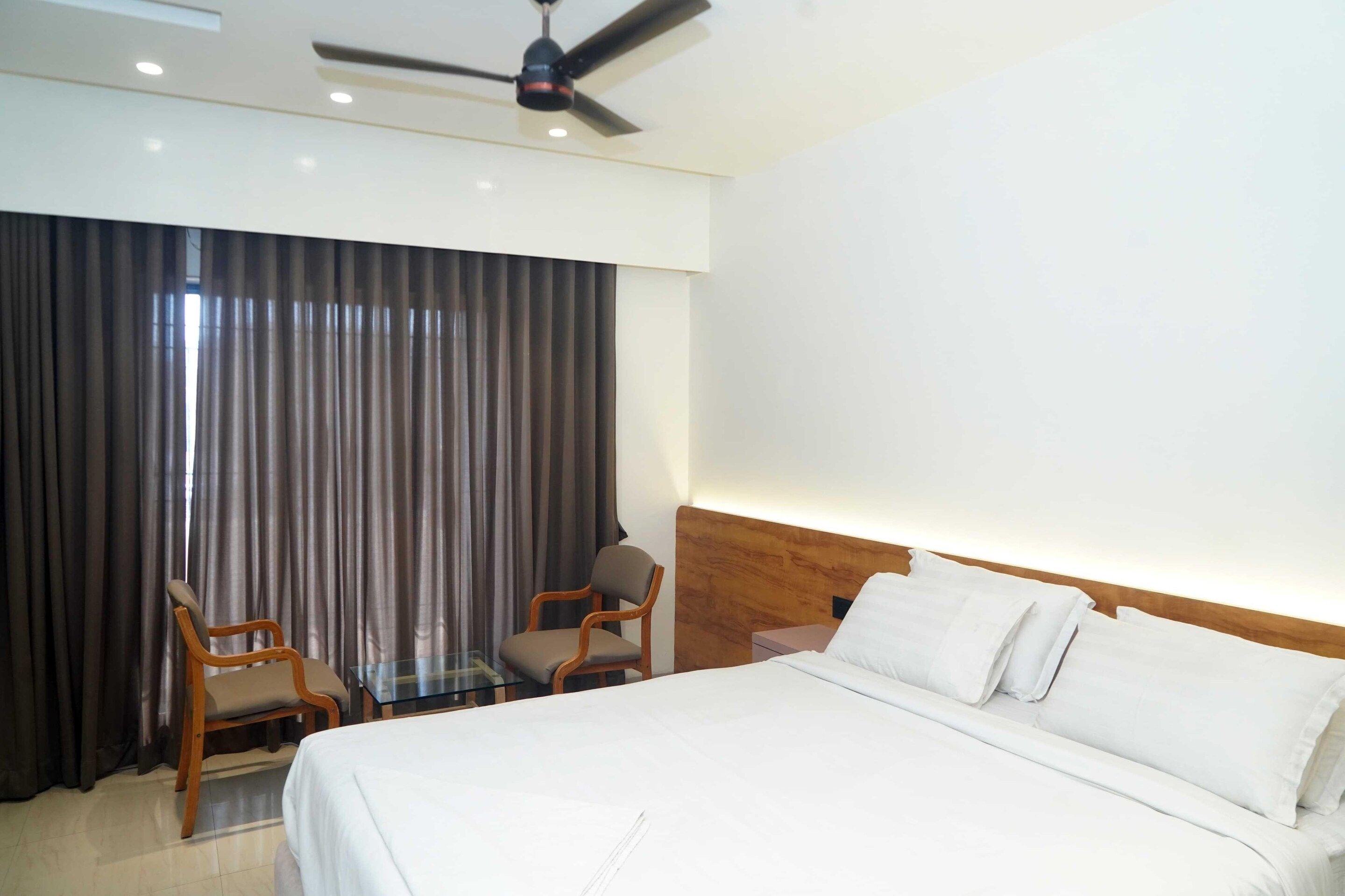 A hotel room with a bed, two chairs, a desk, a ceiling fan, and a large window with curtains.