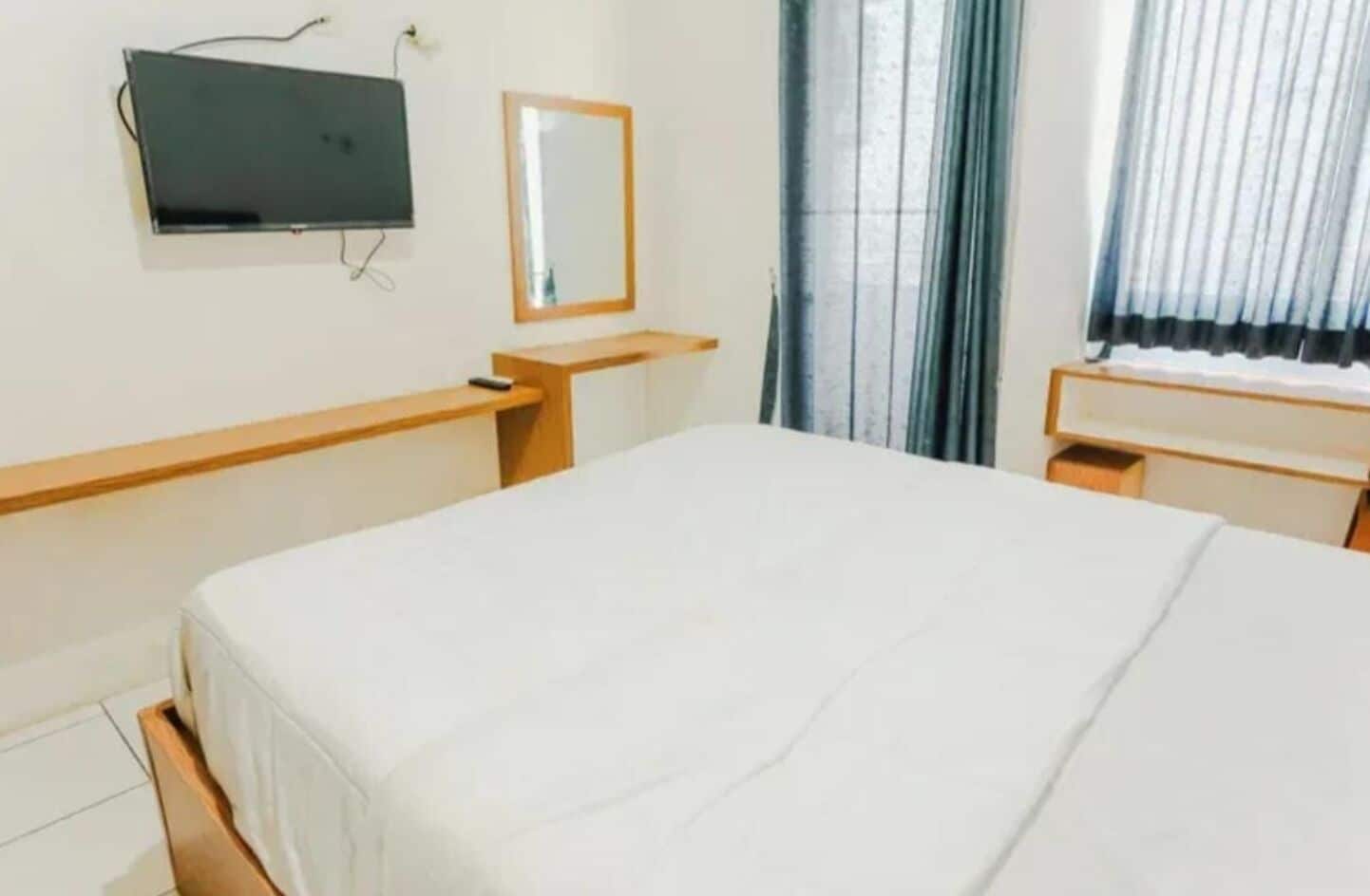 Una habitación de hotel con cama, escritorio, silla, televisor montado en la pared y ventana con cortinas.