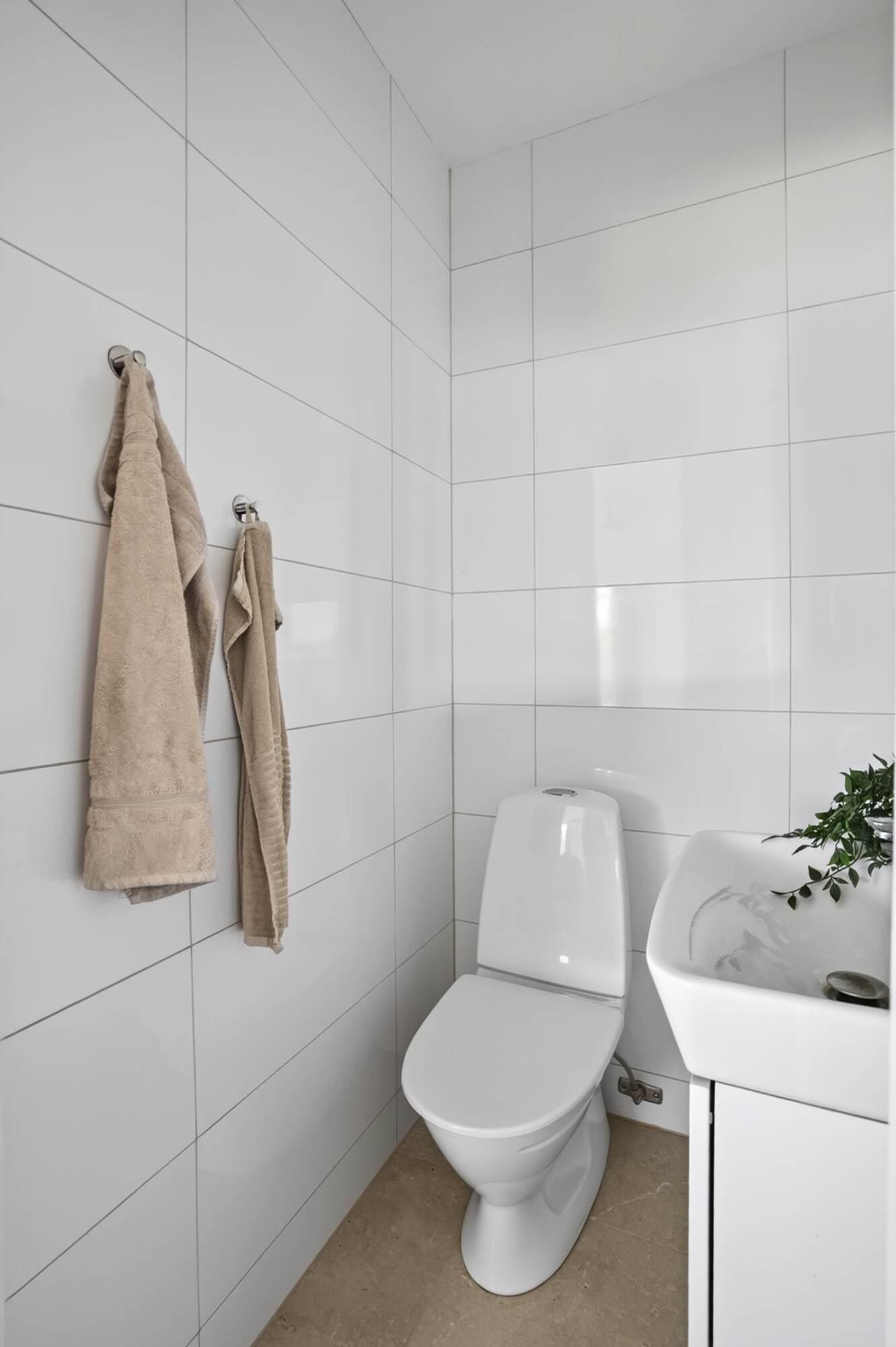 Une salle de bain avec des murs recouverts de carreaux blancs, des toilettes blanches et un lavabo agrémenté d’une plante à côté.