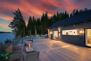 A modern house with a deck overlooking a body of water, surrounded by trees.