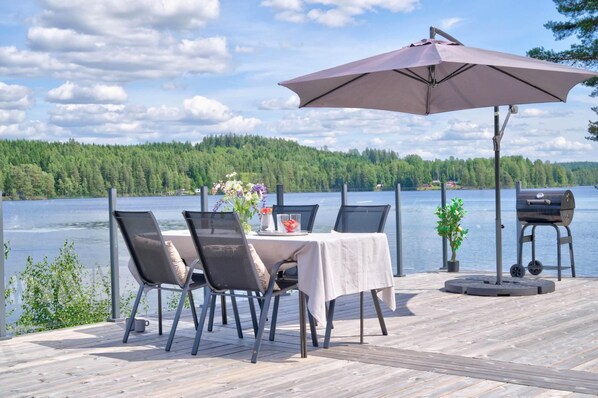 A deck with a table and chairs, a grill, and a view of a lake and forest.