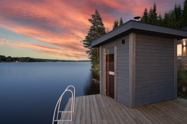A wooden dock with a metal ladder leading to a small cabin by a calm lake at sunset.