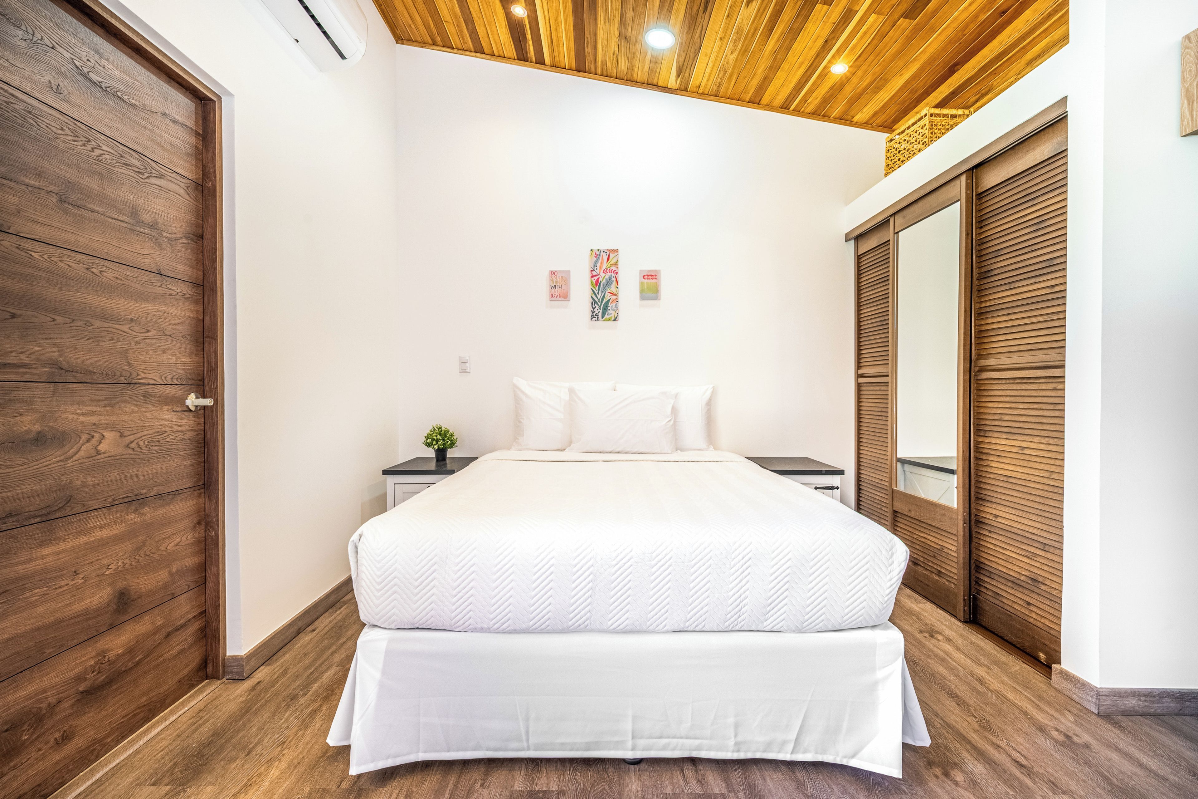 A bedroom with a wooden ceiling, a bed with white bedding, a nightstand with a plant, and a wardrobe with wooden shutters.